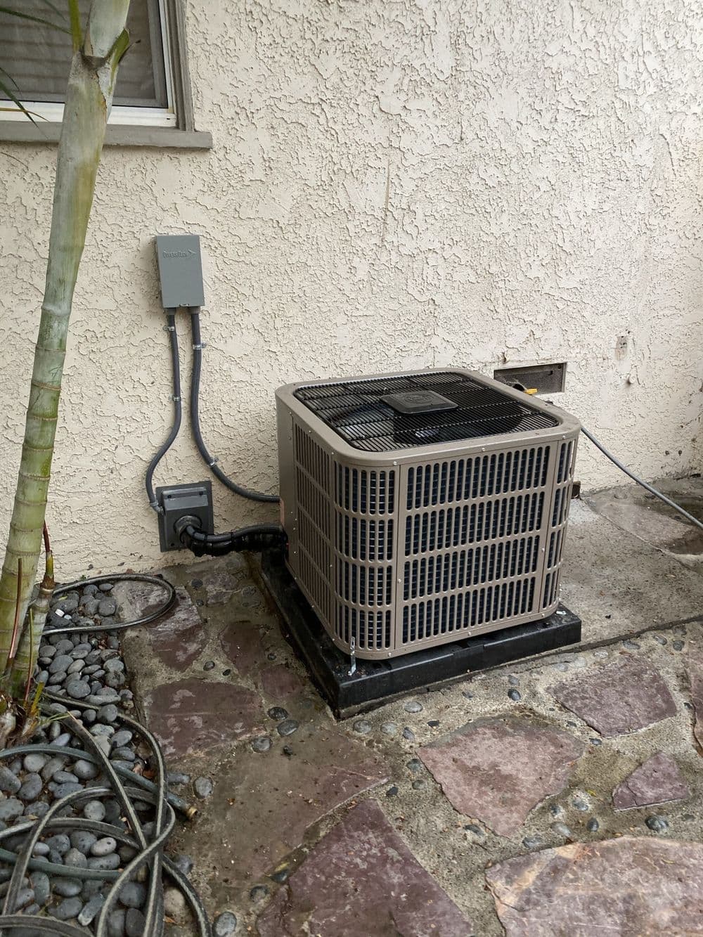 Residential air conditioning unit installed on a stone surface beside a house wall.