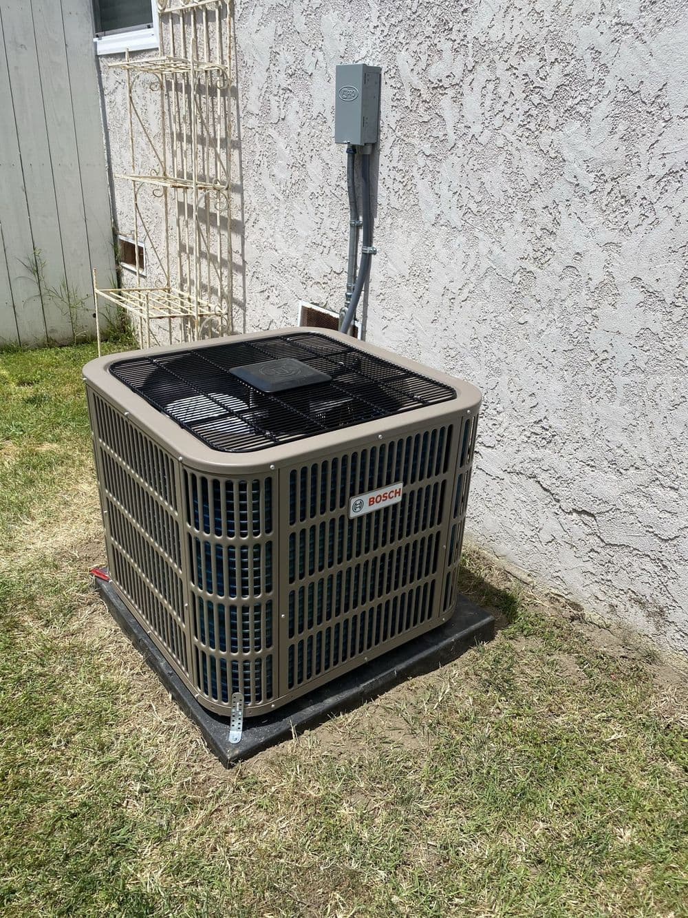 Bosch air conditioning unit installed in yard with grass and wall in background.